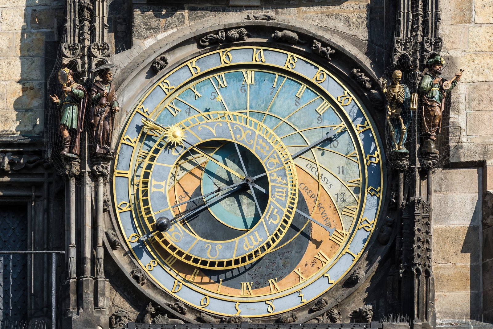 A large round astronomical clock dial in blue, gold and ochre with Roman numerals and zodiac signs, a black sphere on a hand at lower left, painted figures with a mirror at the left and a skeleton and a lute player at the right, on a stone tower wall