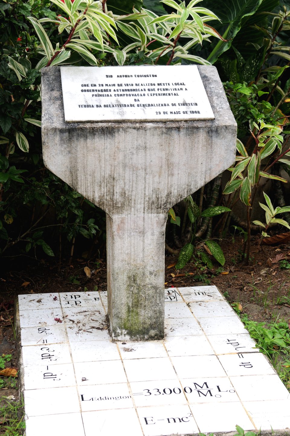 A concrete pillar carrying a memorial plaque, standing in a garden on a tiled base inscribed with physics equations