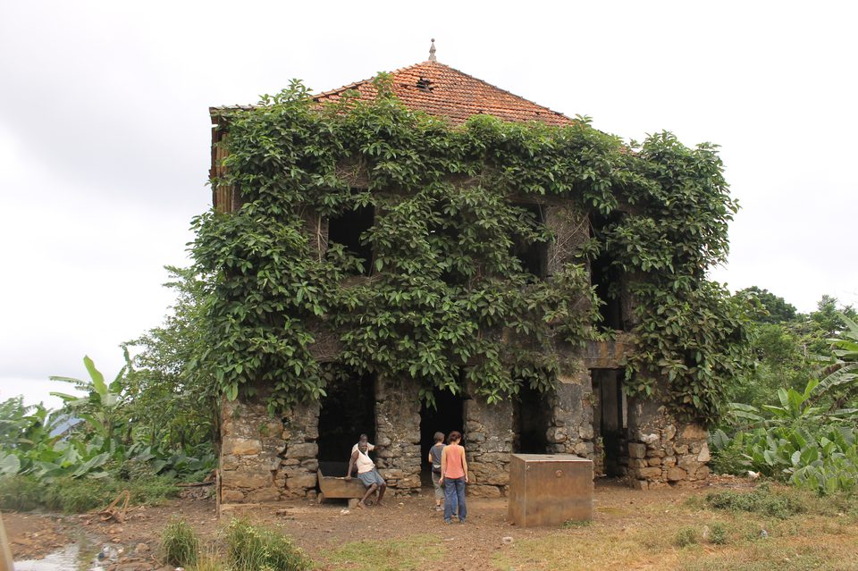 A two-storey stone plantation building almost entirely covered in climbing vines, its tiled roof still intact, two people standing at the arched doorways