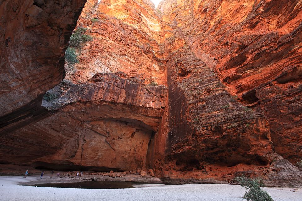 A vast orange rock amphitheatre curving overhead above a pale sandy floor and a still pool, tiny figures at the base