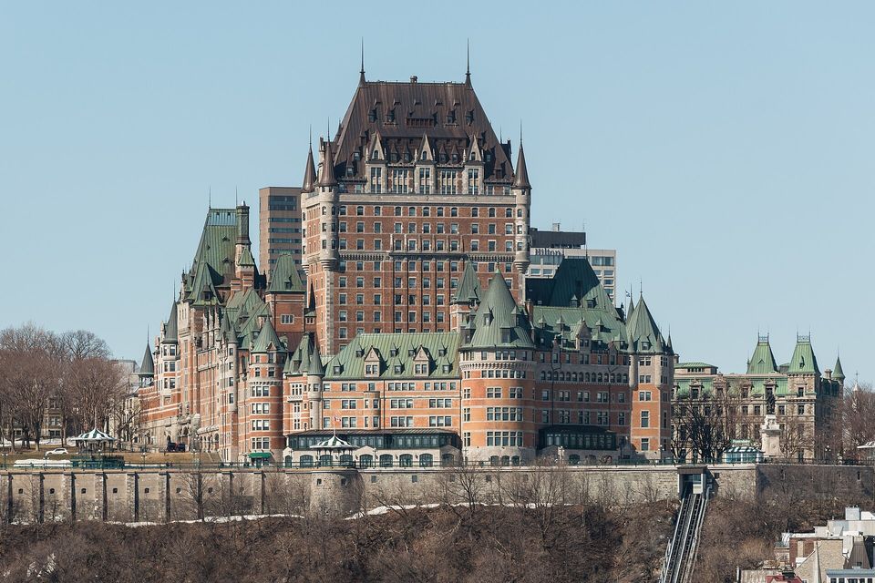 A large brick and green-roofed chateau on a cliff edge above a stone retaining wall and a steep funicular track, bare trees at the left and a clear blue sky