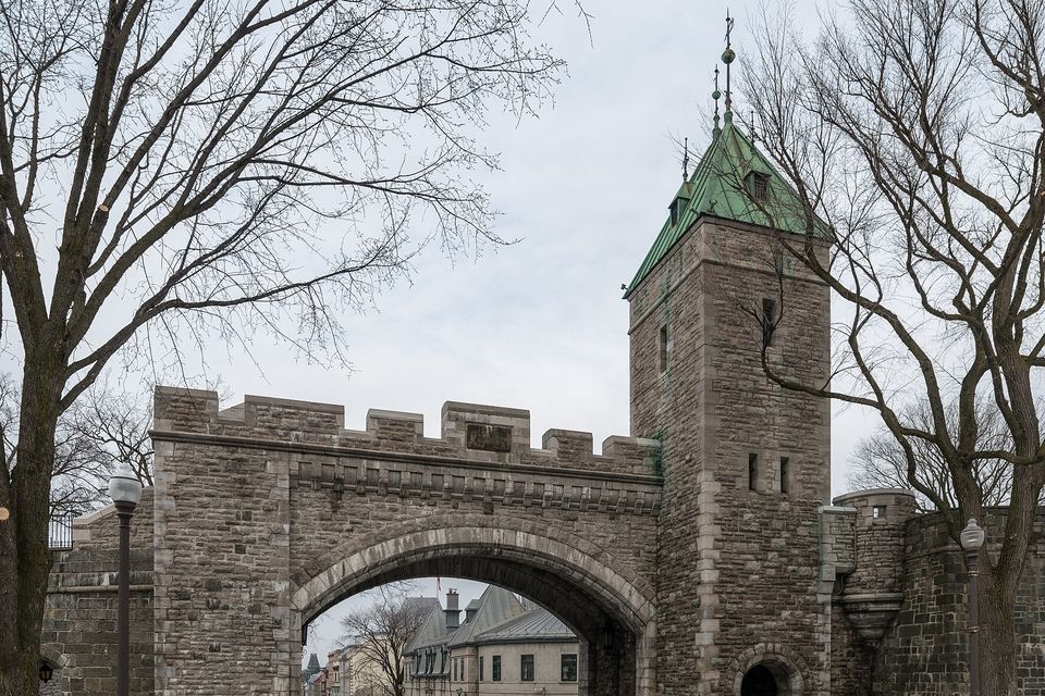 A crenellated stone gateway with a wide arch and a square tower with a green copper roof in the city walls, bare trees on both sides, a street and stone houses seen through the arch