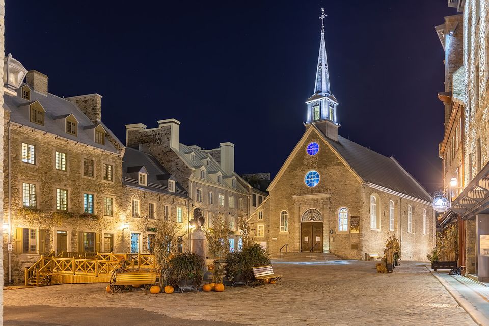 A small cobbled square at night with a grey stone church with a slender lit spire and blue round windows beside three-storey stone houses with warmly lit windows