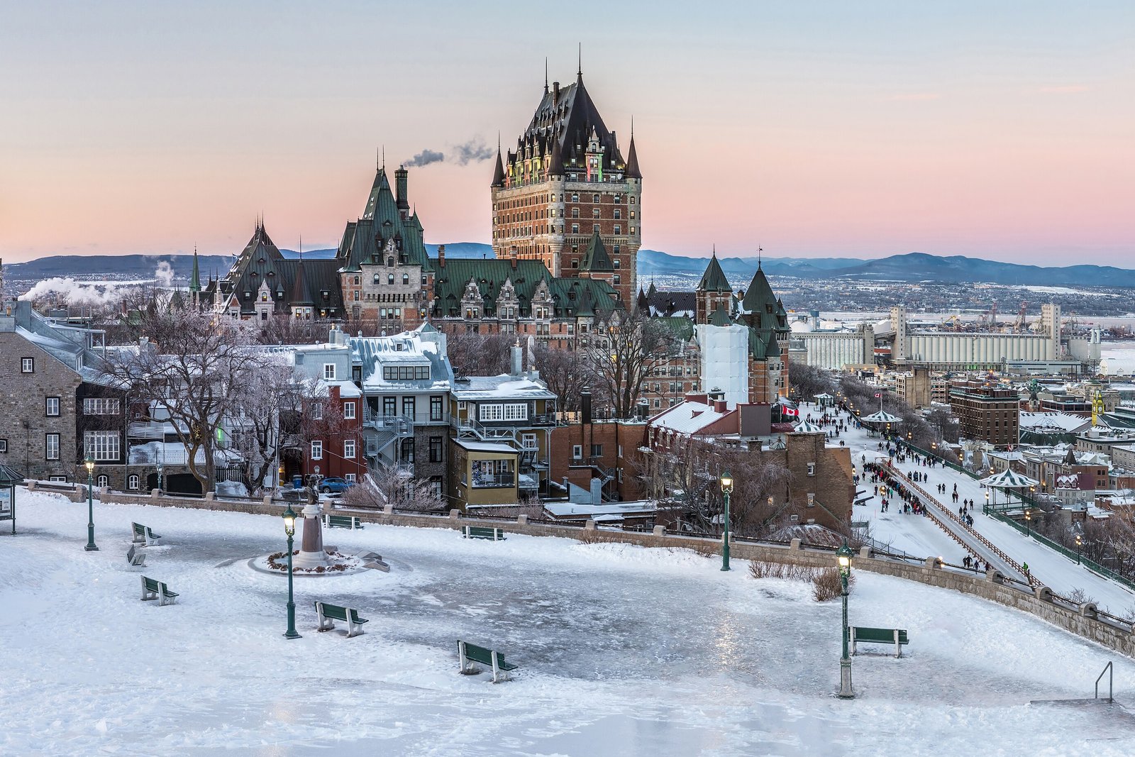 A tall brick chateau with green copper roofs and steep gables at pink dawn above a frozen, snow-covered square with benches and lamp posts, the ice-choked river and far hills beyond