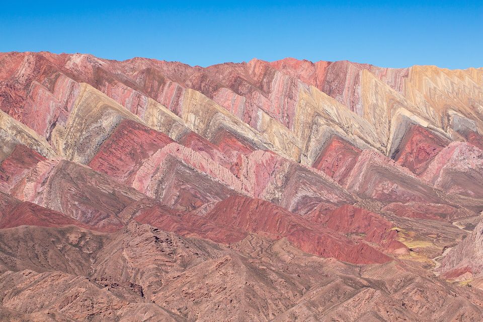 Steep zig-zag mountain ridges striped pink, maroon, ochre and cream under a clear blue sky