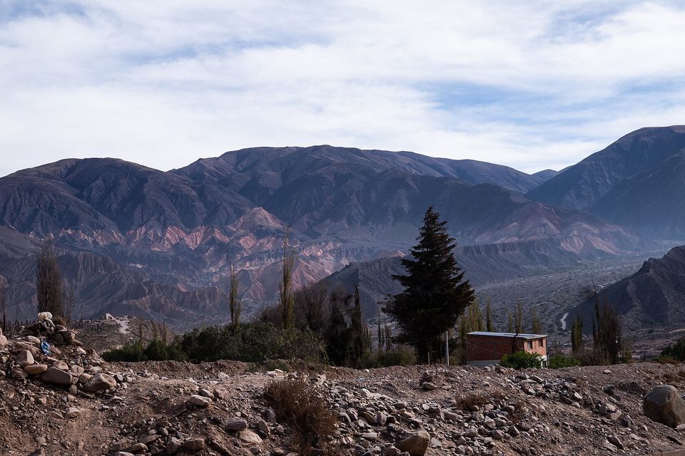 Bare brown mountains with bands of pink and cream rock rise over a stony valley with a dark cypress, poplars and a brick house