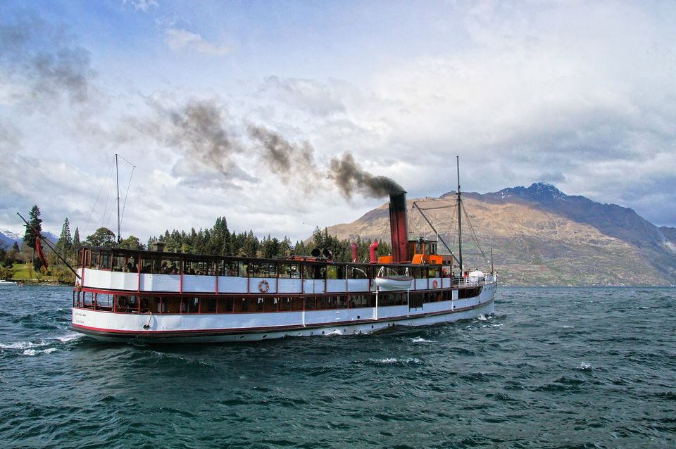 A steamship with a red funnel pouring black smoke on a blue lake below mountains