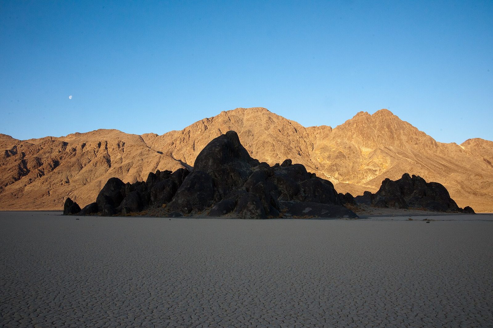 The Grandstand's dark boulder island sits on the cracked white playa, the bare mountains behind lit gold by the first sun, a half moon in a deep-blue sky