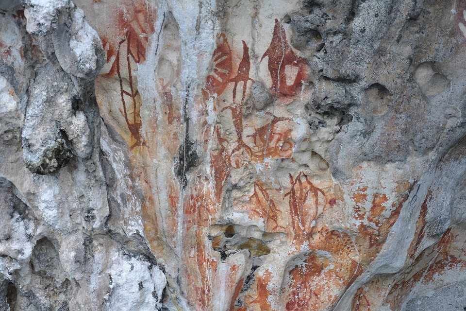 Red-ochre painted figures and hand shapes on a pale, pitted limestone cliff face