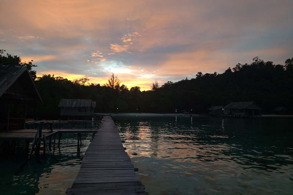 A wooden jetty running out over calm green water toward thatched stilt huts and dark forest under an orange dusk sky