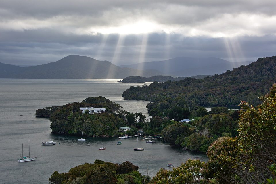 Shafts of light from grey cloud over a bush-covered bay with moored boats and a few houses
