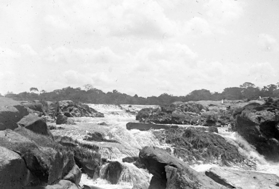 A black-and-white photograph of a broad river tumbling over dark granite ledges in white cascades, a low line of forest on the horizon