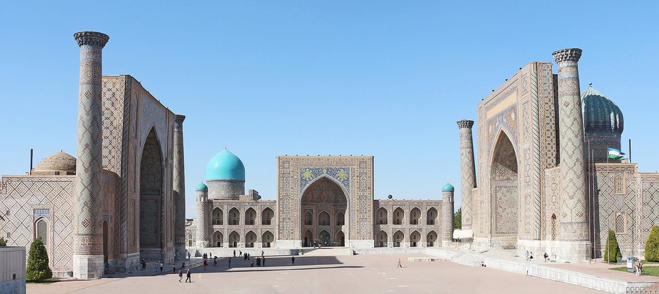 Three tiled madrasas around a wide paved square in clear daylight, with two tall minarets at the outer sides and a turquoise dome behind
