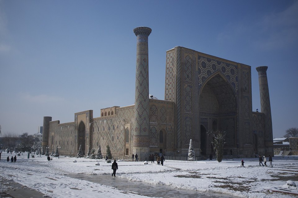 A large tiled madrasa portal between two tall minarets over a snow-covered square, with a few small walkers under a dark blue sky
