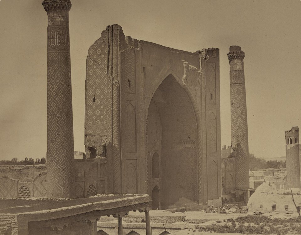 A sepia photograph of a ruined madrasa portal between two tall minarets, with low buildings and bare ground around it