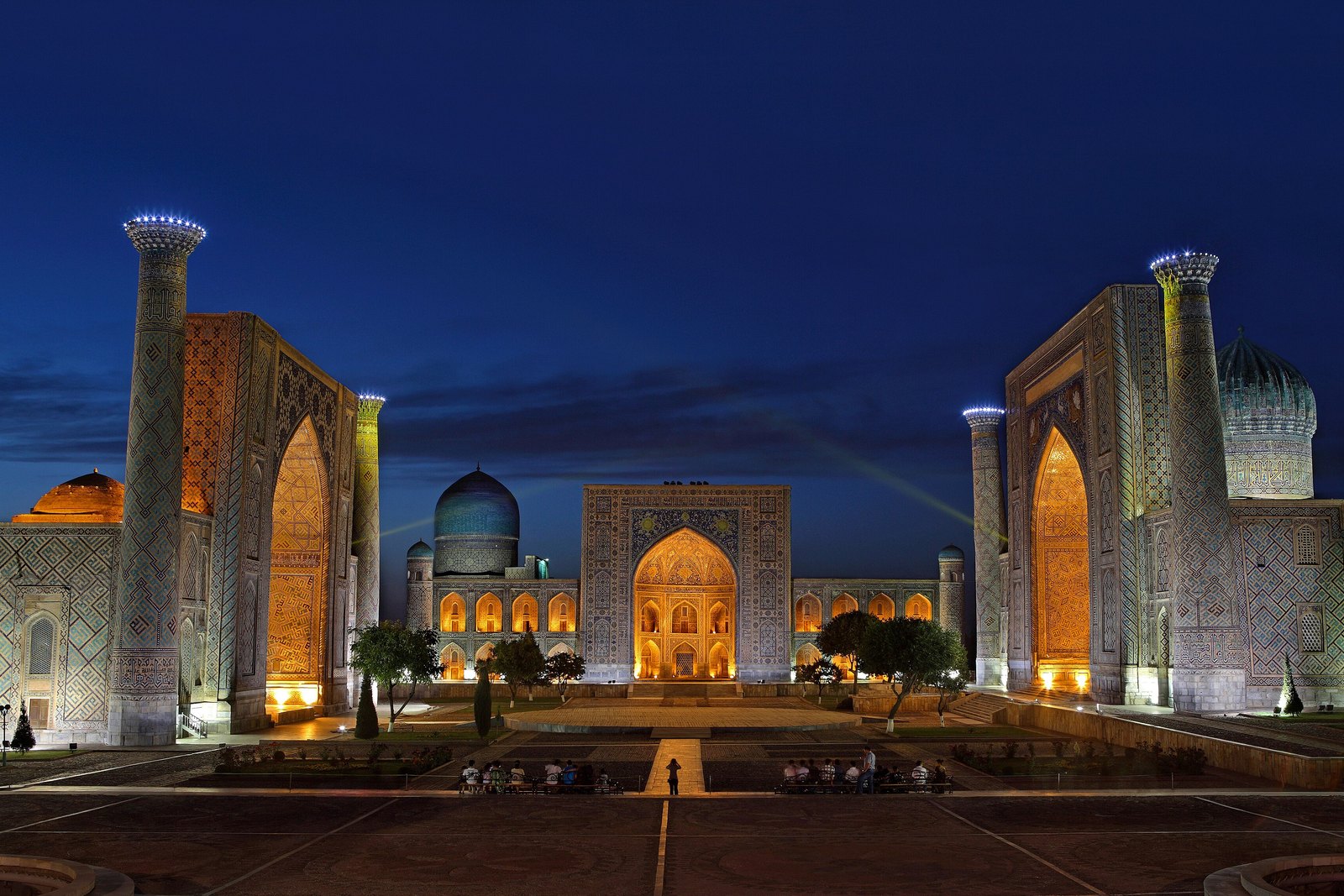Three tiled madrasas with lit portals and tall minarets around a paved square under a deep blue evening sky, a few tiny figures on benches