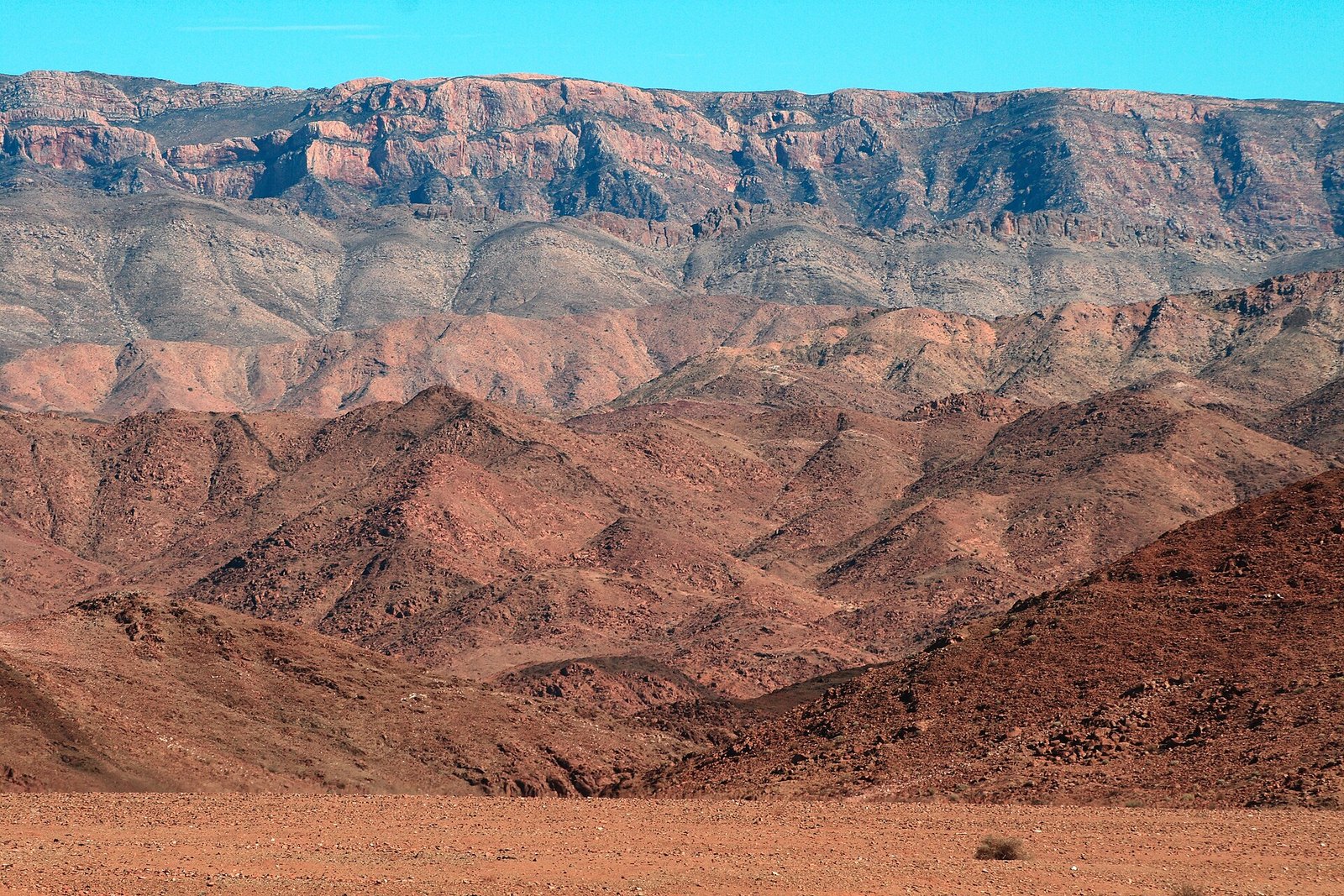 A stack of mountain ridges in rust, rose and slate grey over a red gravel plain