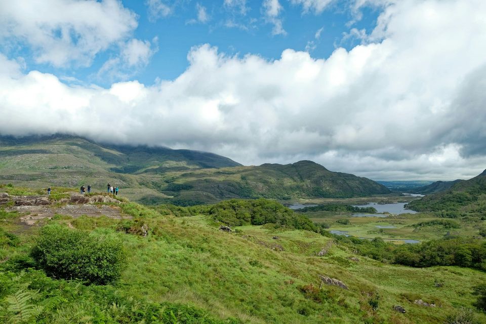A wide valley with a lake under rolling cloud and mountains beyond, a few small walkers at left