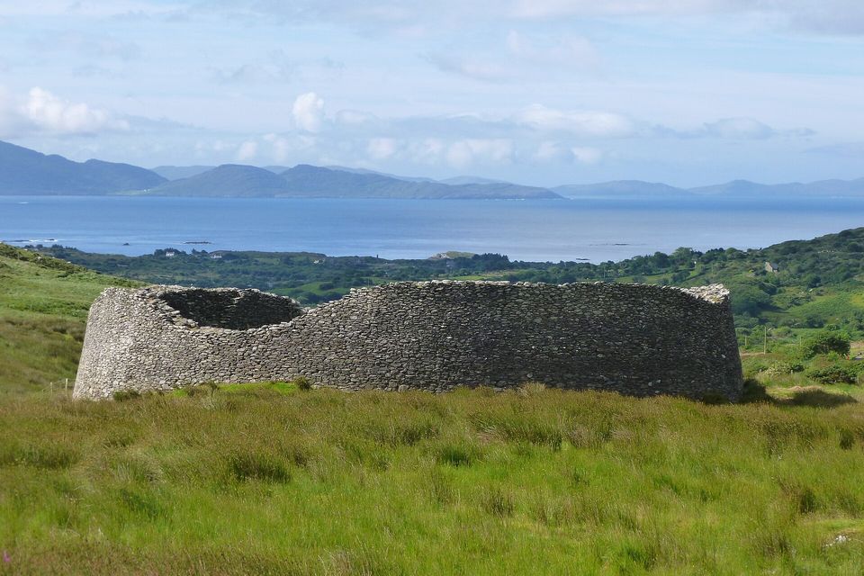 A round dry-stone fort on green pasture with a wide estuary and hills behind under a pale sky
