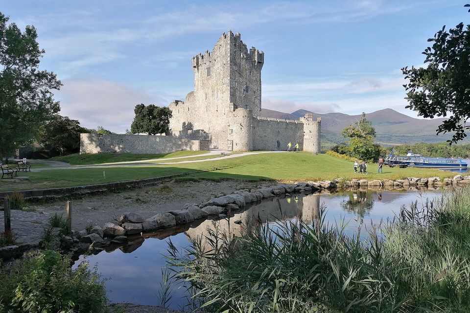 A grey stone tower house with a curtain wall on a lake shore, mirrored in a pool, mountains behind