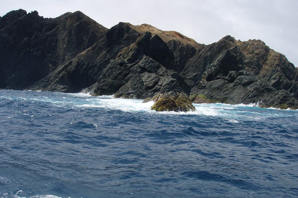 Black layered volcanic cliffs with surf and deep blue sea under a white sky