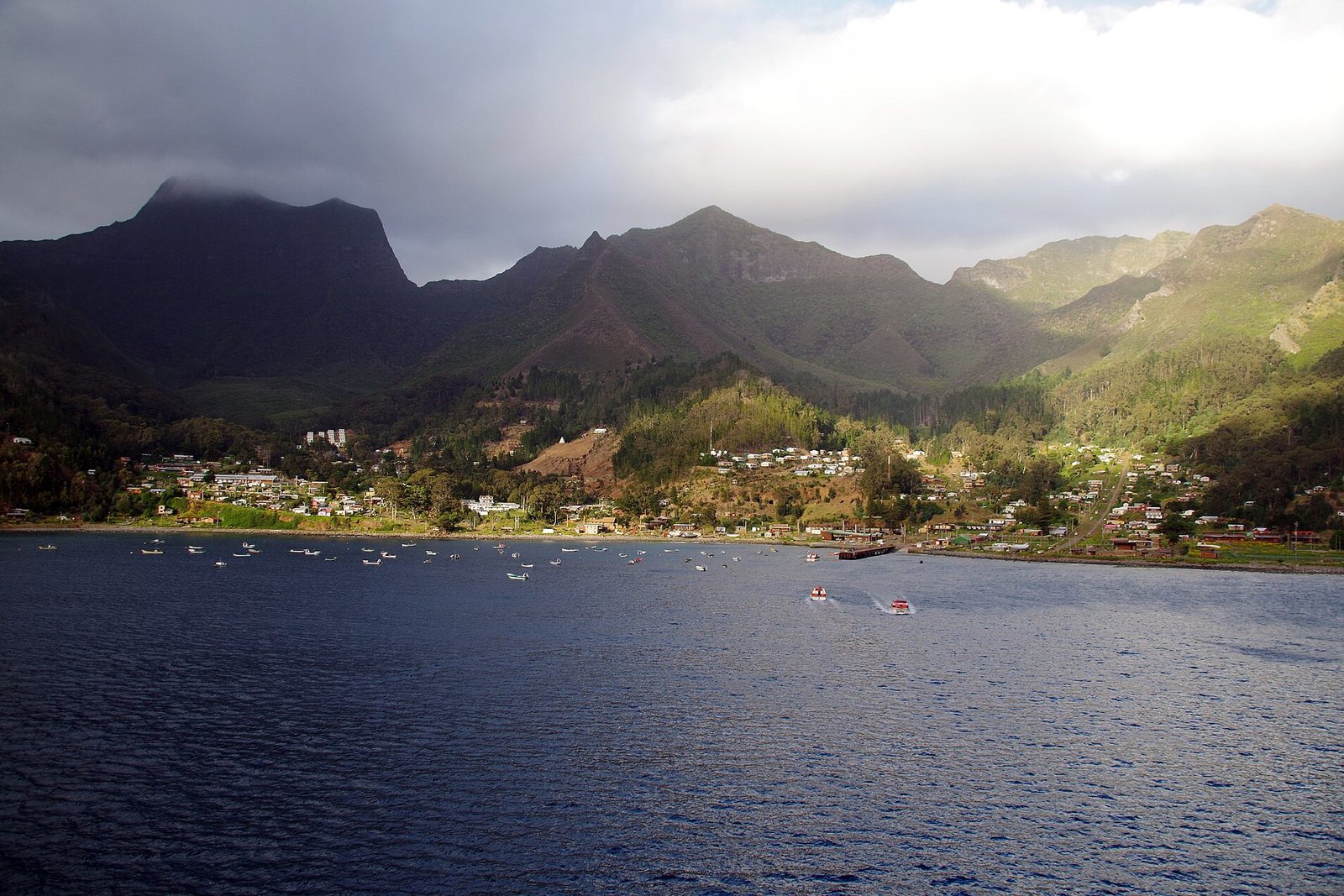 Dark volcanic ridges with a cloud-capped summit above a small town along the shore and deep blue water