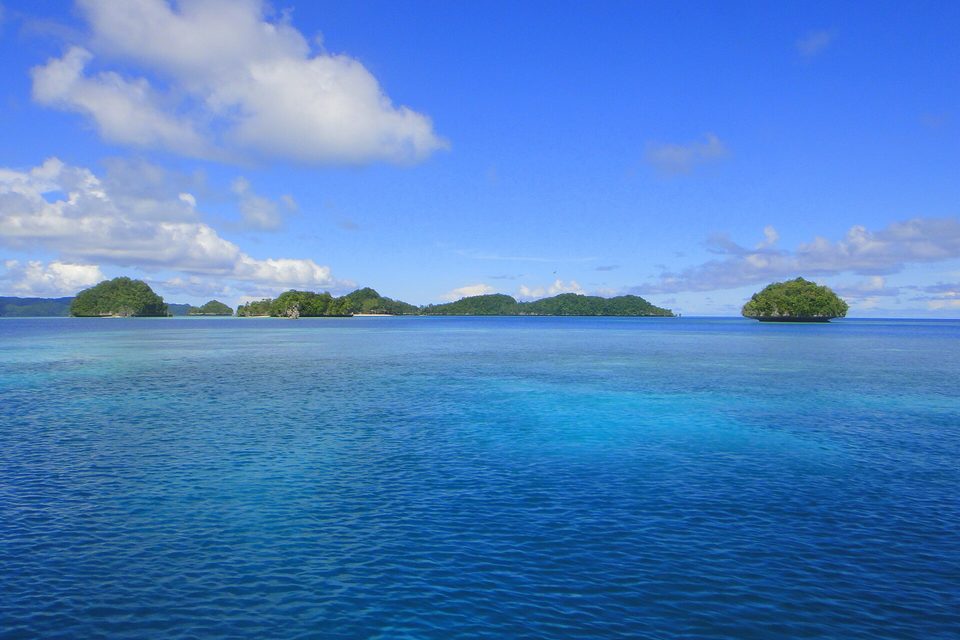 Calm turquoise and deep-blue water in the foreground, low green islets along the horizon and a rounded island at the right, under blue sky and cloud