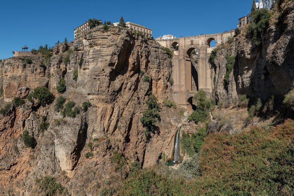 A stone bridge high across a gorge between sheer brown and ochre cliffs, a slim waterfall below and a hotel on the rim at left