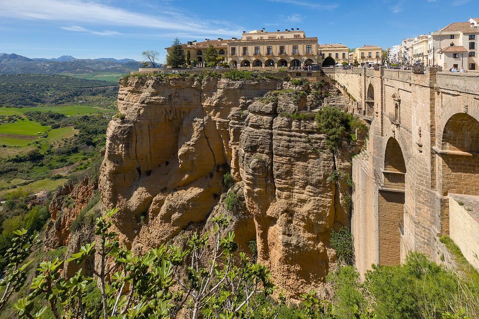 A sandstone cliff falling from the rim of a town, an arcaded building above, a green plain and distant hills at left and the edge of a stone bridge at right
