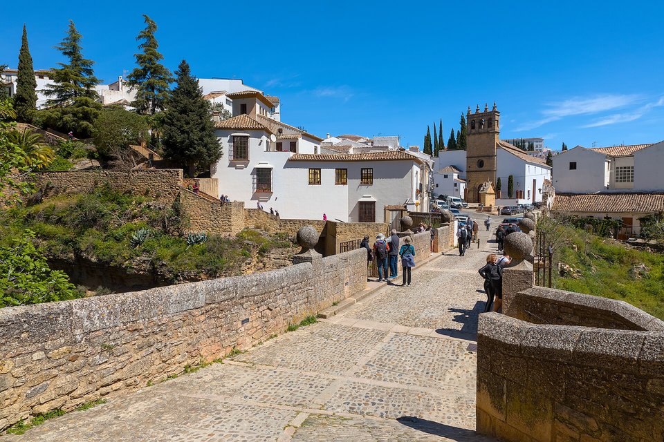 A cobbled stone bridge with low parapets leading to whitewashed houses, cypresses and a square stone church tower under a deep blue sky, a few walkers on the bridge