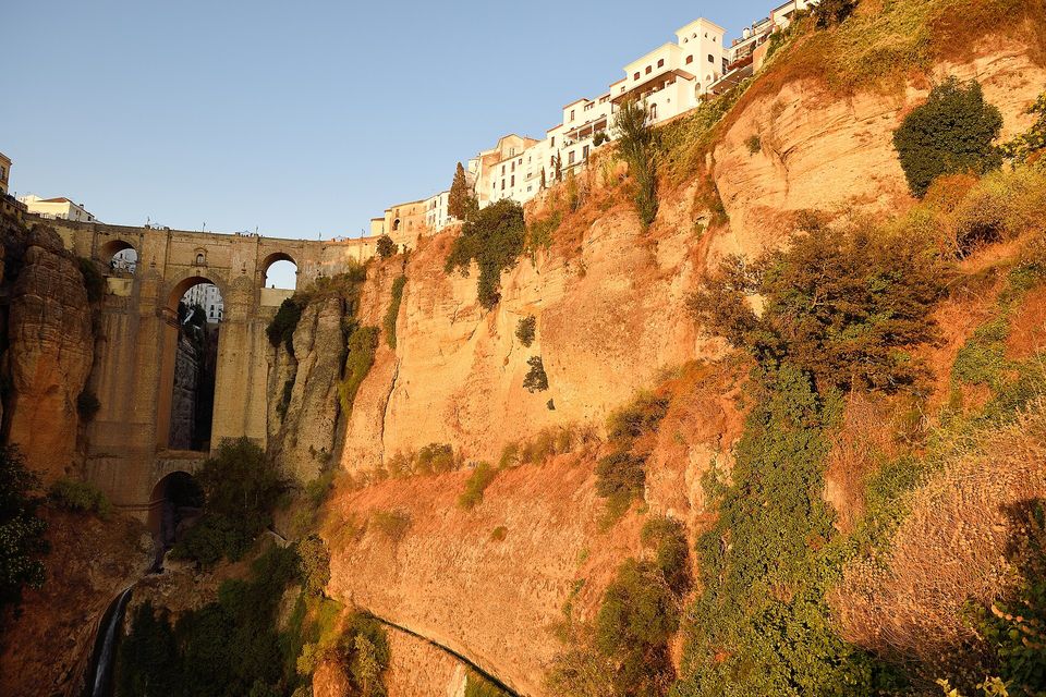 A stone bridge with tall arches at the left and a sheer orange cliff catching the low sun, white houses along its top and a pale sky