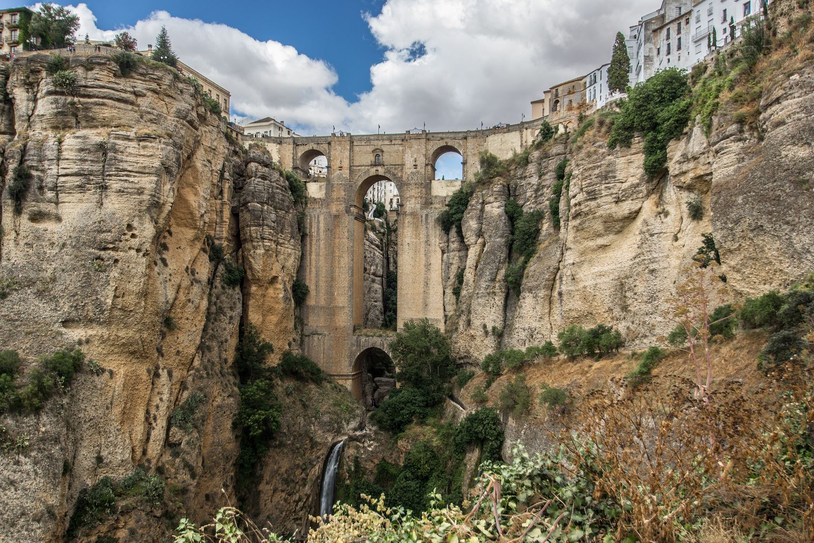 A tall stone bridge with three arches spanning a narrow gorge between pale layered cliffs, white houses along the rim and a waterfall at the foot, under a blue sky with clouds