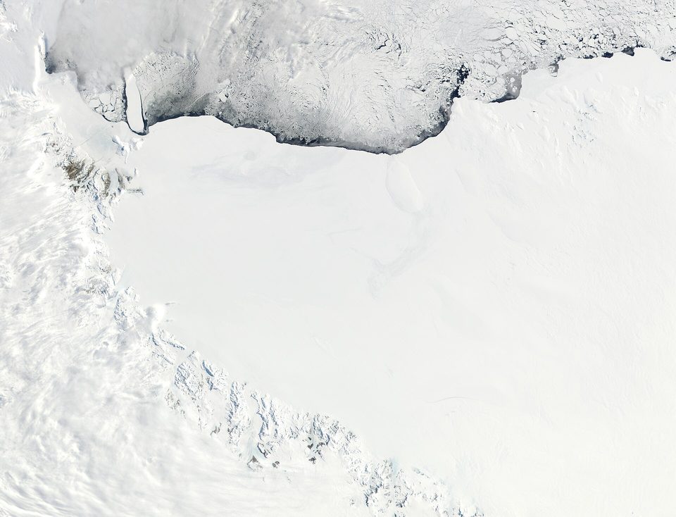 A satellite view of a vast white ice sheet with cracked, darker broken sea ice along its upper edge and rugged mountain ice at the left