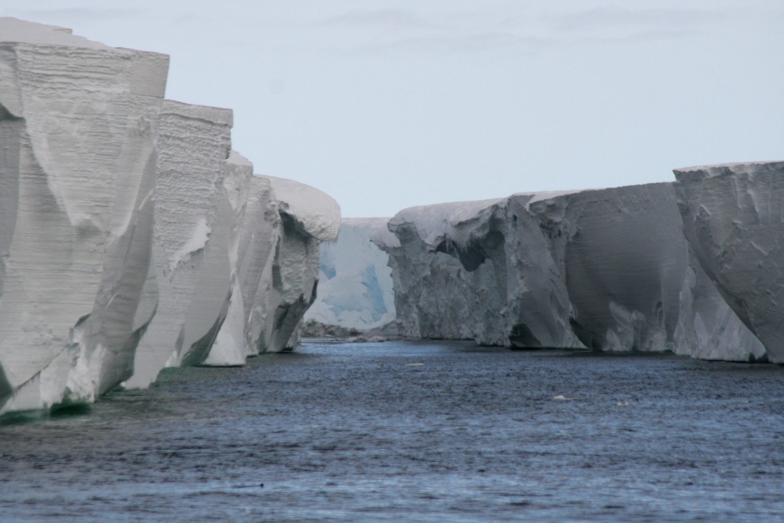 Two pale grey ice cliffs with flat tops and sculpted, streaked faces rising from dark grey water either side of a narrow inlet, blue ice in the gap, pale sky above