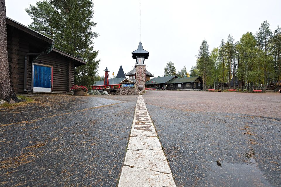 A white painted line lettered ARCTIC CIRCLE running along a paved square toward a stone marker post and log buildings