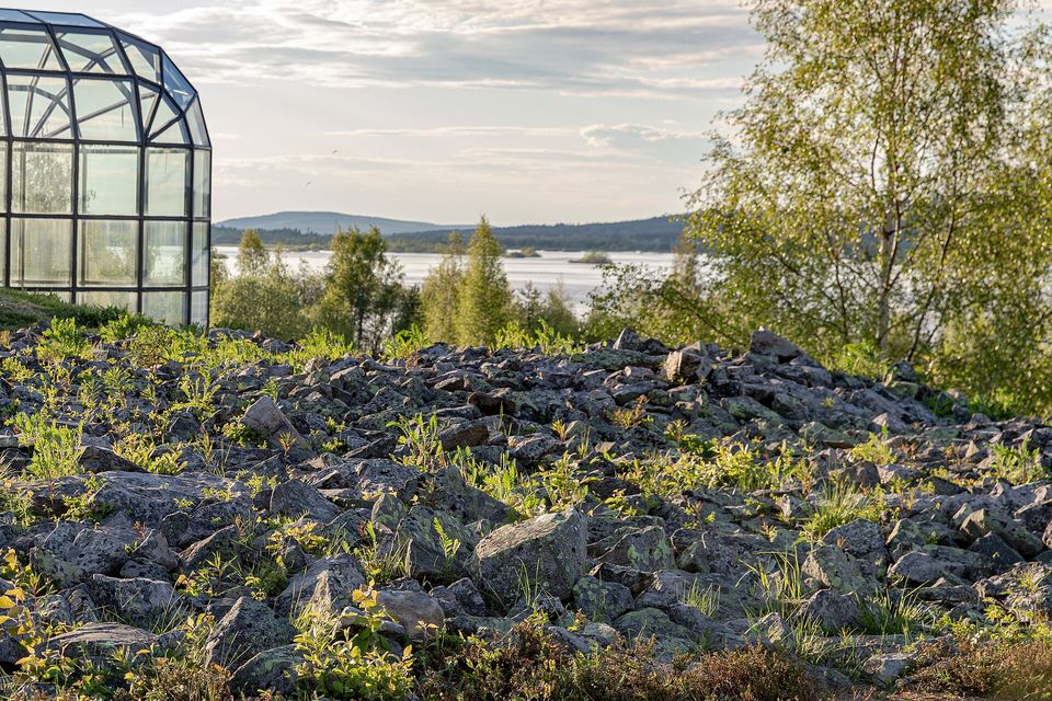 A glass gallery at the left above a field of grey boulders with small plants, a wide river and a low hill beyond under a pale evening sky