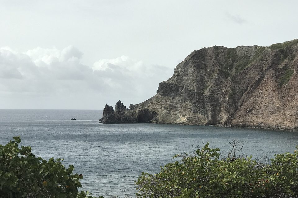 Dark volcanic cliffs dropping into the sea along a bay, with a point beyond under a grey sky