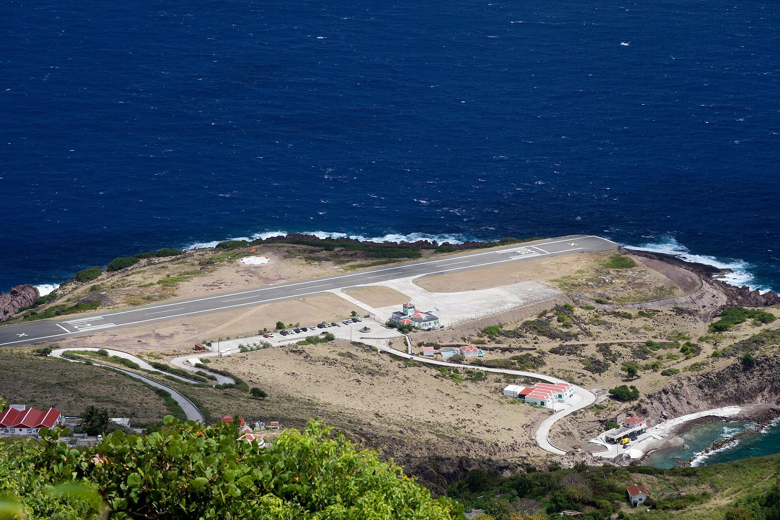 A short runway on a cliff above a deep blue sea, with red-roofed terminal buildings, a switchback road and surf on the rocks