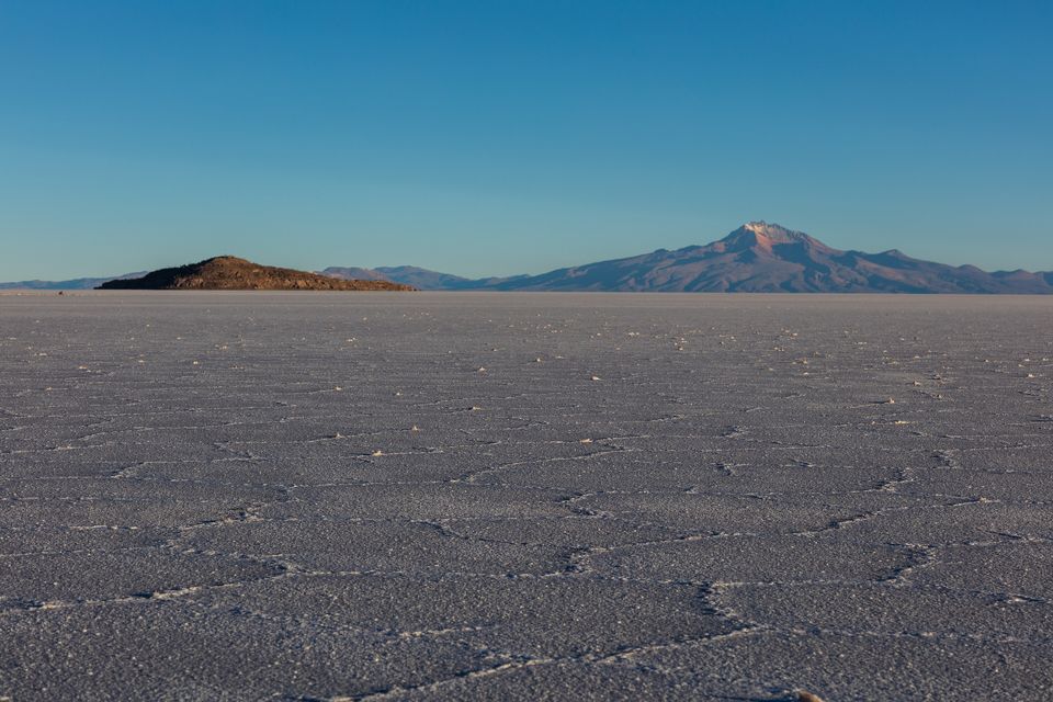A grey salt crust cracked into polygons stretching to a low hill and a distant mountain under a teal sky