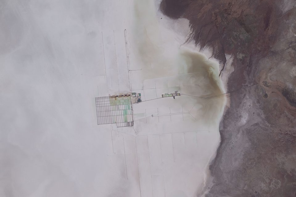 An astronaut photograph of the edge of a white salt flat with rectangular lithium evaporation ponds and an industrial plant