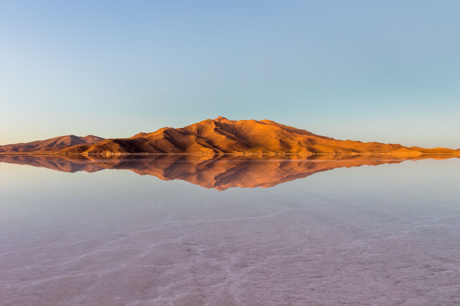 The flooded salt flat as a perfect mirror at sunrise: a low ochre hill in golden light reflected in still water under a pale blue sky