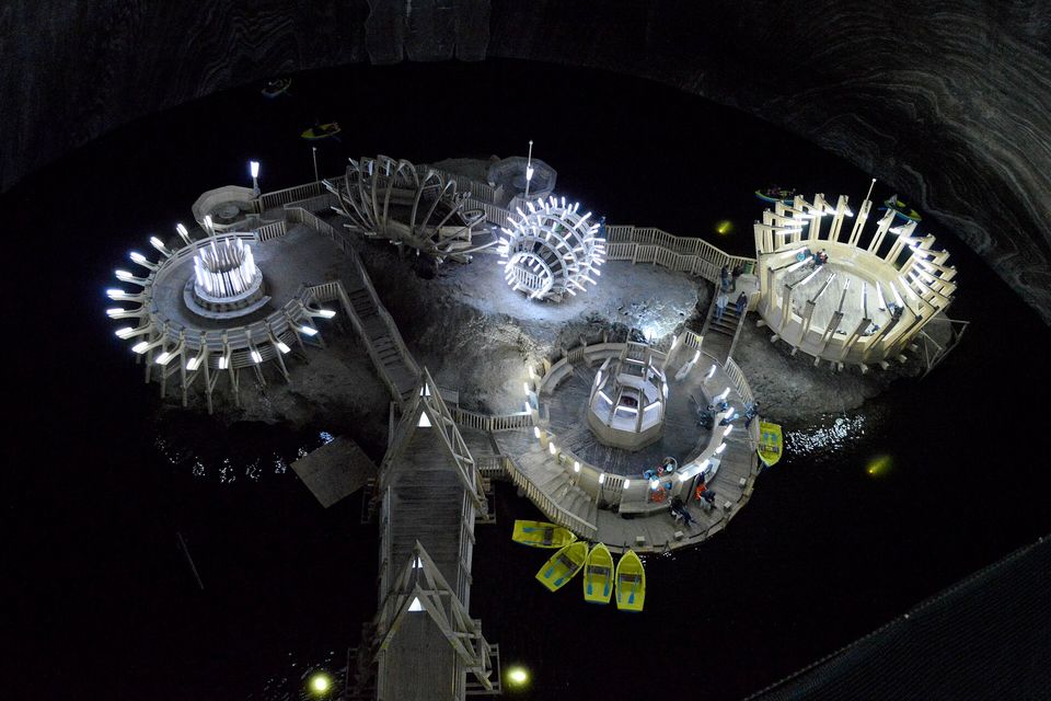 Seen from high above, a salt island on a black underground lake carries lit timber pavilions and walkways, yellow rowing boats moored at its edge