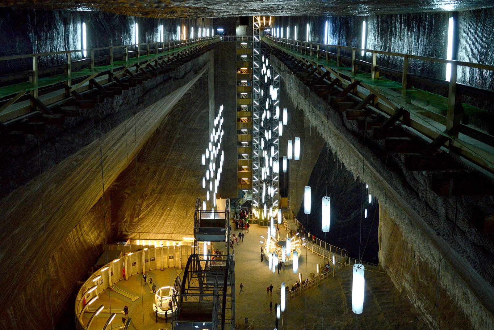 Looking down a vast hall of banded salt walls, two timber walkways run toward a lift tower and strings of white lamps hang into the void, a Ferris wheel and amphitheatre on the floor far below