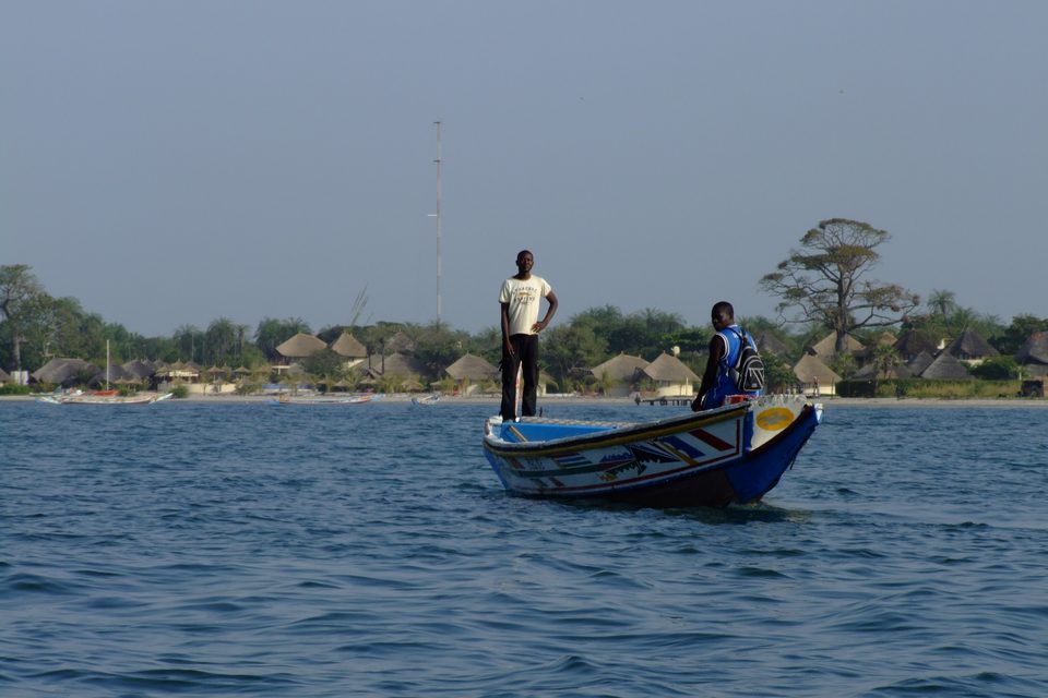 Two men standing in a brightly painted pirogue on open water, a village of thatched roofs and a baobab on the far shore