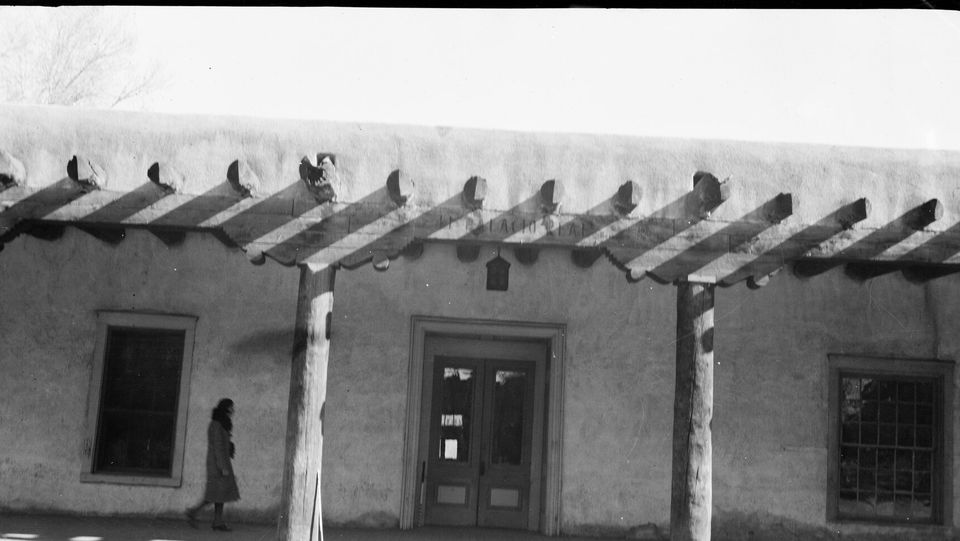 A long adobe front with a timber-posted portal and projecting roof beams, a single passer-by in the shade, in black and white