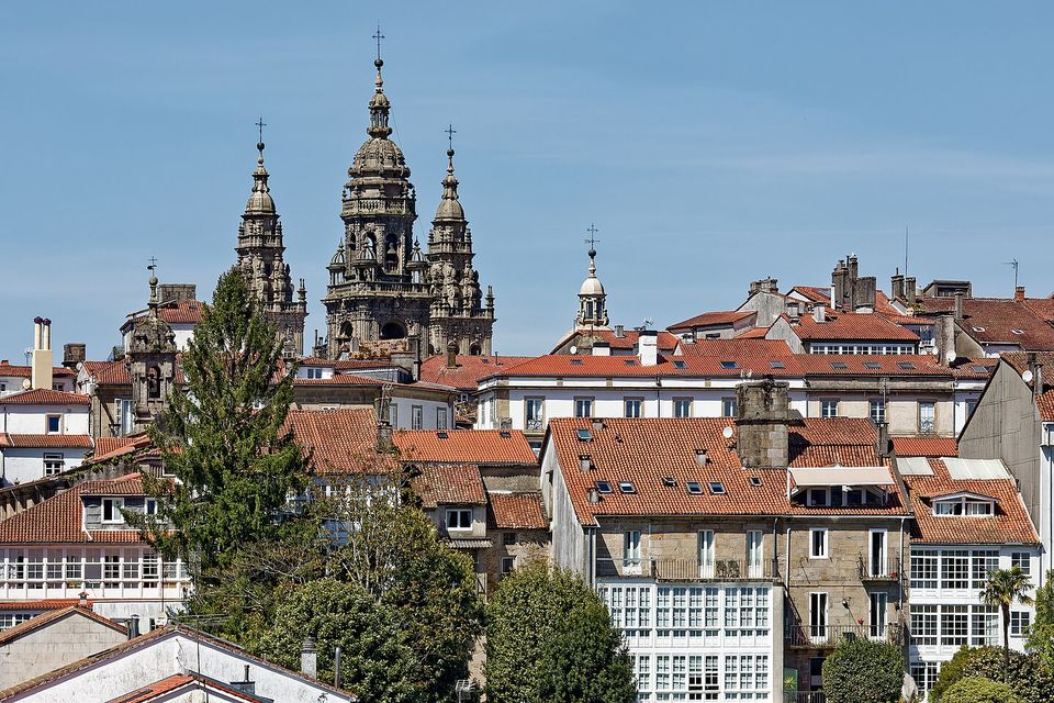 The twin towers of a cathedral seen over red roofs and white galleried houses, with trees and a shop sign in the foreground