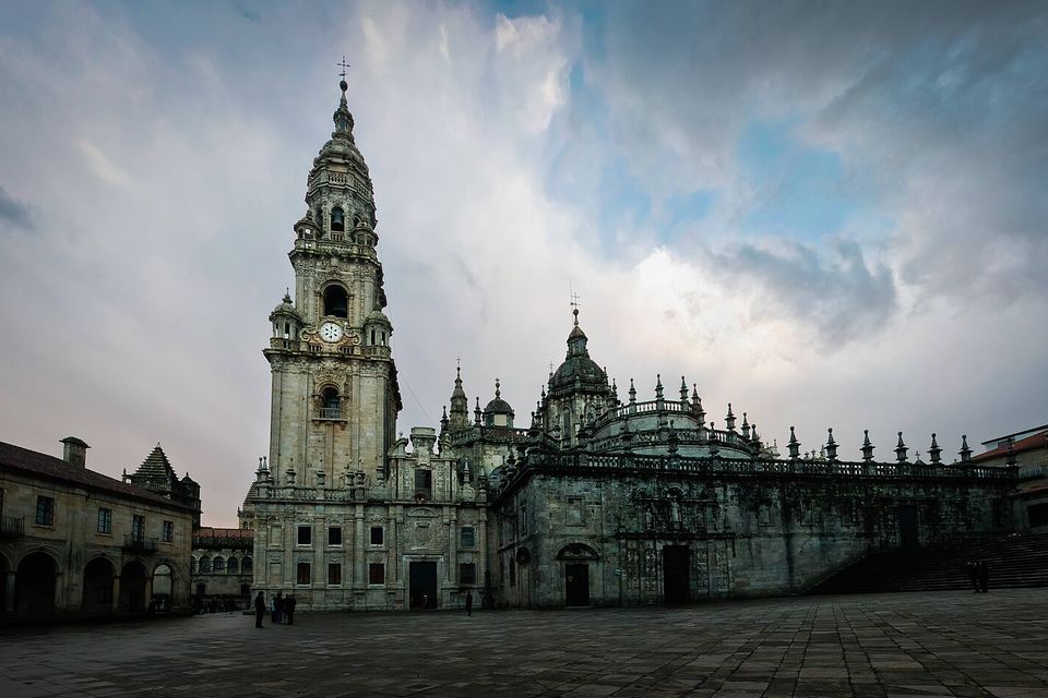 A tall clock tower and the carved stone east end of a cathedral above an empty paved square, under a dark stormy sky