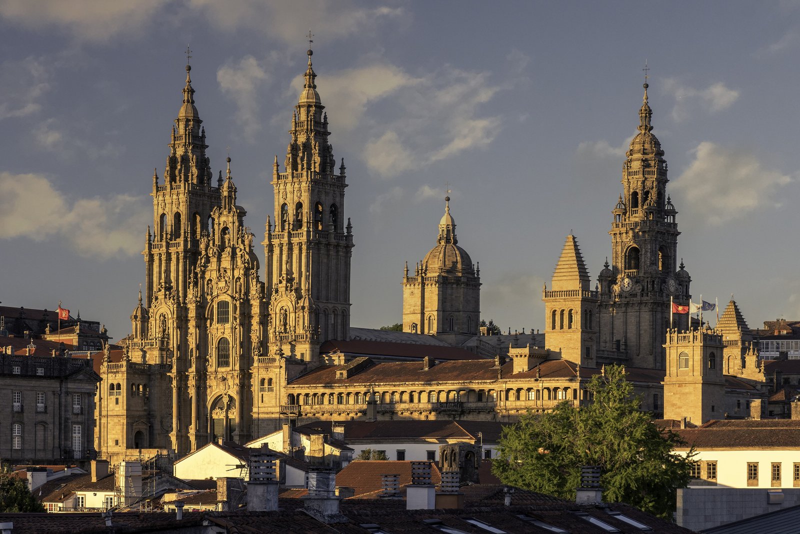 Two tall ornate baroque towers, a dome and a clock tower of golden stone lit by low sun above dark tiled roofs, under a blue sky with broken cloud