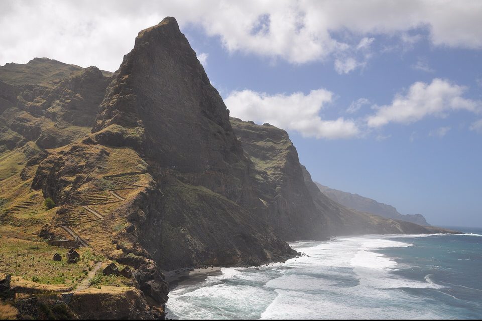 A steep dark headland with stone terraces and small huts on its flank drops to white surf under a blue sky