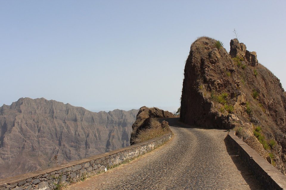 A cobbled road with low stone walls curves past a rock pinnacle, with jagged brown ridges beyond under a pale sky